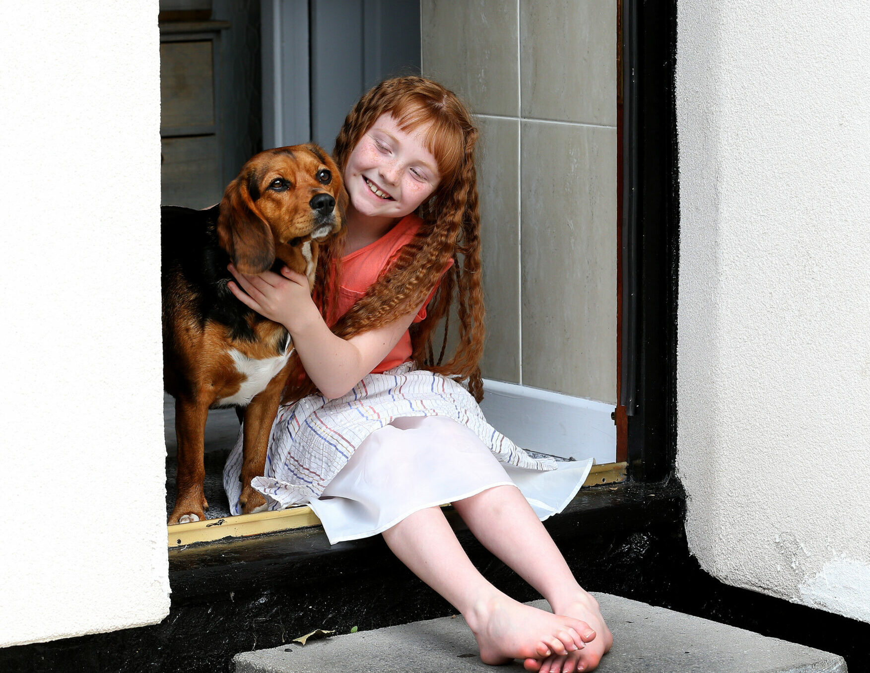 Young girl happily sits at the step of her house, cuddling a large dog.
